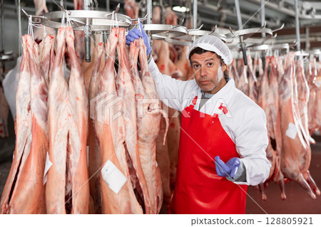 Male slaughterhouse worker showing mutton carcass in meat storage 128805921