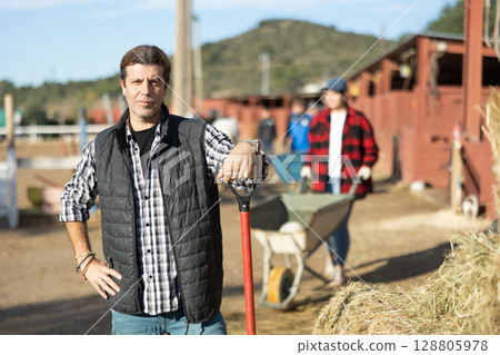 Skilled adult man stables worker in plaid shirt looking at camera while holding working tool on horse ranch in spring Skilled adult man stables worker in plaid shirt looking at camera while holding working tool on horse ranch in spring 128805978