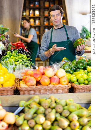 Male seller holding green bell peppers standing in fruit and vegetable section of supermarket Male seller holding green bell peppers standing in fruit and vegetable section of supermarket 128805981