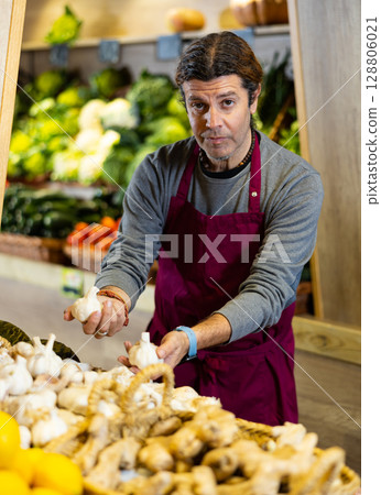 Male seller holding garlic standing in fruit and vegetable section of supermarket 128806021