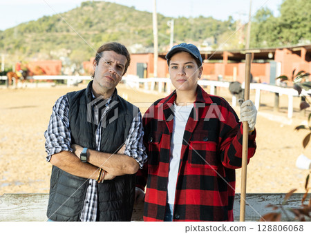 Experienced man and woman in plaid shirts looking at camera while holding working tool on horse ranch in spring Experienced man and woman in plaid shirts looking at camera while holding working tool on horse ranch in spring 128806068
