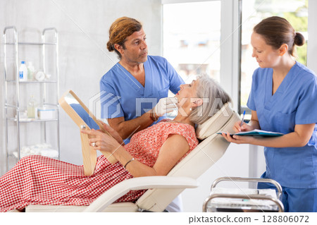 Man dermatologist examines skin of senior womans face, female nurse records data in client card Man dermatologist examines skin of senior womans face, female nurse records data in client card 128806072
