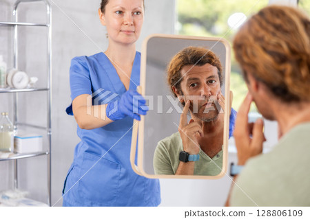 Man carefully examines his face with mirror after cosmetic surgery in clinic 128806109