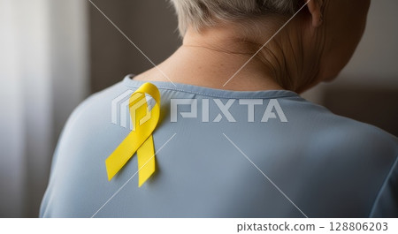 Close-up back view of European woman wearing light blue shirt with bright yellow awareness ribbon pinned on shoulder blade area Close-up back view of European woman wearing light blue shirt with bright yellow awareness ribbon pinned on shoulder blade area 128806203