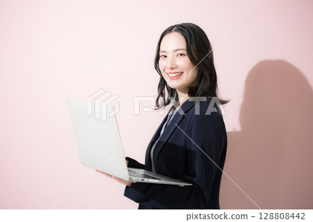 Upper body of a young cute business woman standing in a suit holding a laptop and looking at the camera, business, etc. on a pink background 128808442