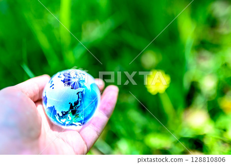 A crystal globe held over a flower blooming in the grass. Ecological image. Light blue, dark blue. A crystal globe held over a flower blooming in the grass. Ecological image. Light blue, dark blue. 128810806