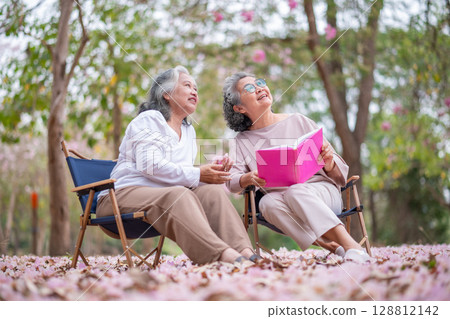 Elderly women under tree adorned pink blossoms, reading book heartwarming and inspiring representation of senior women embracing technology, nature lifestyle. Elderly women under tree adorned pink blossoms, reading book heartwarming and inspiring representation of senior women embracing technology, nature lifestyle. 128812142