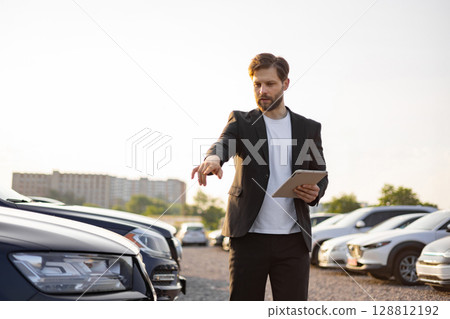A car salesman in a suit points to a vehicle while holding a tablet in a parking lot on a sunny day, demonstrating a car sale. 128812192