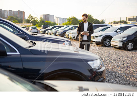 A businessman in a suit uses a tablet while standing in a car dealership, surrounded by rows of new cars on a sunny day. 128812198