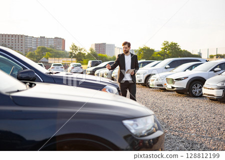 A car salesman in a suit gestures toward a row of cars, holding a tablet, set against a parking lot backdrop. 128812199