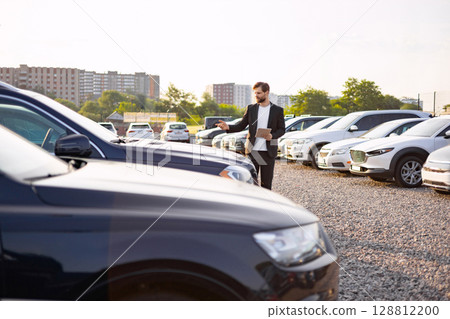 A car salesman points towards a vehicle on a sunny day at a car dealership. Many new cars are on display. A car salesman points towards a vehicle on a sunny day at a car dealership. Many new cars are on display. 128812200