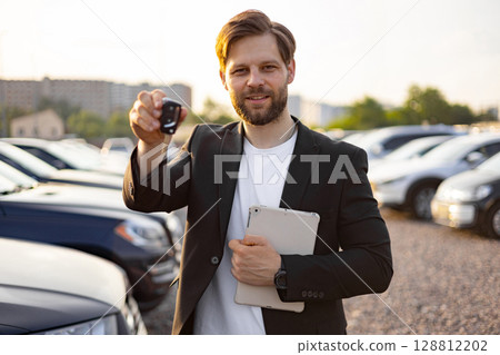 A car salesman, holding a car key and tablet, smiles towards the camera in a car dealership. Cars are visible in the background. 128812202
