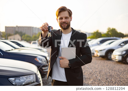 A smiling car salesman holding keys, standing in a car lot, surrounded by various vehicles, suggesting car buying or ownership. 128812204