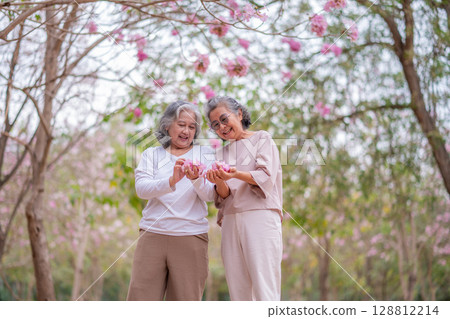 Elderly women with gray hair enjoying a beautiful moment together outdoors, surrounded by blooming pink flowers expressions reflect deep friendship and happiness relaxation with nature. 128812214