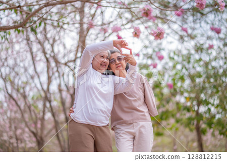 Elderly women with gray hair enjoying a beautiful moment together outdoors, surrounded by blooming pink flowers expressions reflect deep friendship and happiness relaxation with nature. 128812215