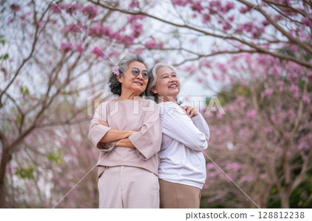 Elderly women with gray hair enjoying a beautiful moment together outdoors, surrounded by blooming pink flowers expressions reflect deep friendship and happiness relaxation with nature. Elderly women with gray hair enjoying a beautiful moment together outdoors, surrounded by blooming pink flowers expressions reflect deep friendship and happiness relaxation with nature. 128812238