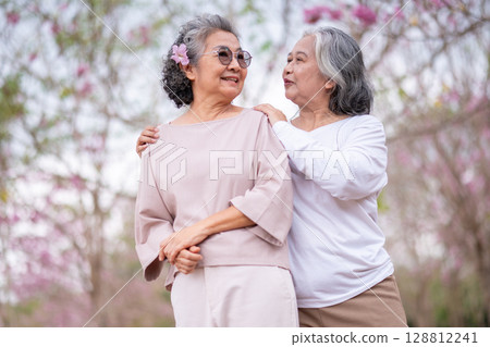 Elderly women with gray hair enjoying a beautiful moment together outdoors, surrounded by blooming pink flowers expressions reflect deep friendship and happiness relaxation with nature. 128812241