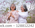 Elderly women with gray hair enjoying a beautiful moment together outdoors, surrounded by blooming pink flowers expressions reflect deep friendship and happiness relaxation with nature. 128812242