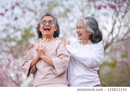 Elderly women with gray hair enjoying a beautiful moment together outdoors, surrounded by blooming pink flowers expressions reflect deep friendship and happiness relaxation with nature. 128812242