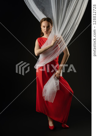 Woman in a flowing red dress poses with sheer fabric in a studio setting, expressing emotion and elegance during a photoshoot 128812728