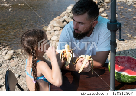 Father and daughter enjoy a sunny picnic by the river, sharing bananas and watermelon. 128812807