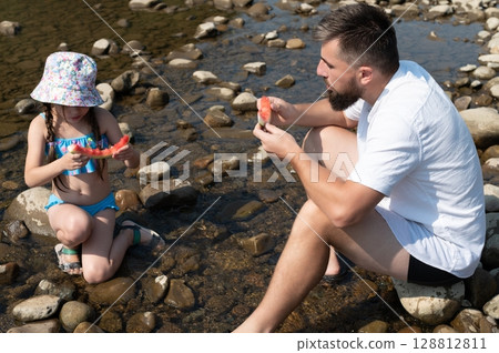 A father and daughter enjoy a refreshing watermelon snack by a shallow river on a sunny day. A father and daughter enjoy a refreshing watermelon snack by a shallow river on a sunny day. 128812811