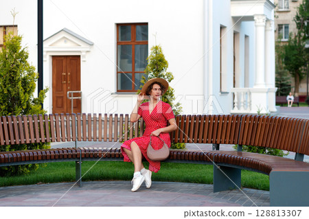 Woman with handbag adjusts hat while sitting on city bench. 128813307