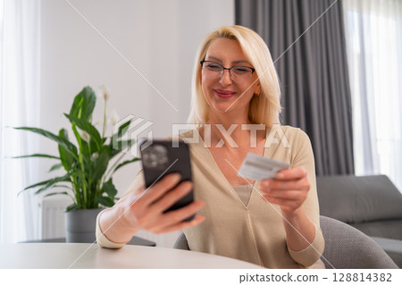 Smiling middle-aged blonde woman using her mobile phone to shop online and pay with a credit card. Concept of modern technology, e-commerce, and easy online payments at home. 128814382