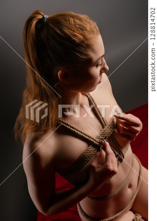 Woman engaged in shibari art with ropes and lingerie against moody backdrop 128814702