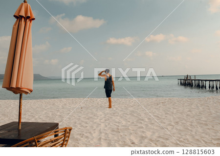 A lone person walking on the sandy beach with an umbrella and wicker chair on the foreground and the ocean in the background. Koh Rong Sanloem, Sihanoukville, Cambodia 128815603