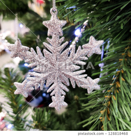 Close-up of a white artificial snowflake with glitter on a Christmas tree branch. Festive photo of Christmas decor. Close-up of a white artificial snowflake with glitter on a Christmas tree branch. Festive photo of Christmas decor. 128818468