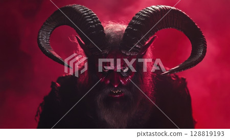 A menacing horned demon figure with glowing eyes and sharp teeth emerges from deep red smoke A menacing horned demon figure with glowing eyes and sharp teeth emerges from deep red smoke 128819193