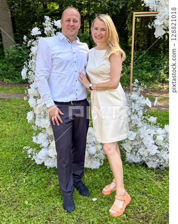 miling couple poses at a stylish outdoor event in front of a floral wedding backdrop. Dressed elegantly, they radiate happiness in a green garden setting. 128821018