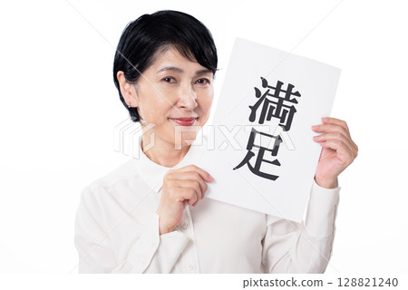 A senior woman wearing a white shirt smiling and holding a piece of paper with the word "Satisfied" written on it 128821240
