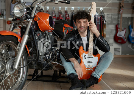 A man holding a guitar stands beside a motorcycle in a vibrant music store setting 128821609