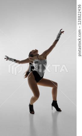Dancer in silver leotard striking dynamic pose in high heels against white background 128822436