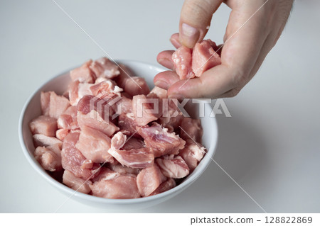 A human hand lifts fresh raw diced meat from a white bowl, representing healthy protein sources, essential ingredients for home cooking, and balanced meal preparation 128822869