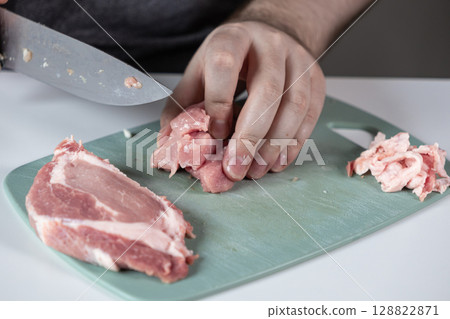 Person slicing raw pork meat with a knife on a green cutting board, preparing ingredients for a healthy home-cooked meal in the kitchen Person slicing raw pork meat with a knife on a green cutting board, preparing ingredients for a healthy home-cooked meal in the kitchen 128822871