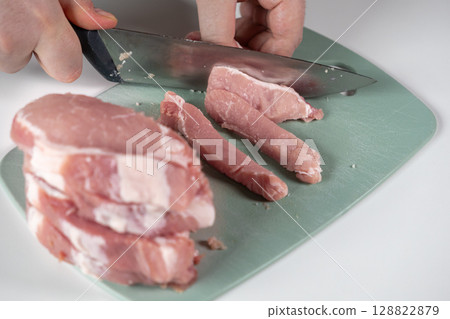 Hands carefully preparing fresh raw pork meat on a green cutting board with a sharp knife, signifying nutritious home cooking, healthy lifestyle, and culinary skill 128822879