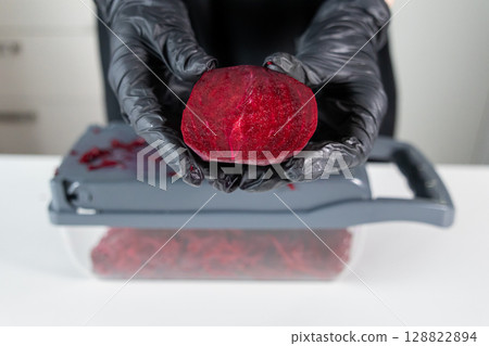 Hands in black protective gloves hold a vibrant red beetroot half over a kitchen grater, demonstrating healthy food preparation and kitchen safety 128822894