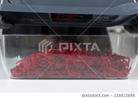 Vibrantly shredded beetroots cascade into a clear glass container from a kitchen food processor, promoting healthy cooking and efficient meal preparation 128822898
