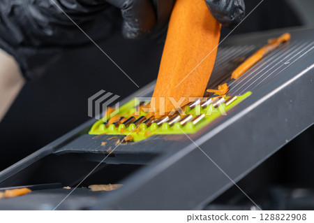 Hand in black glove expertly grating a vibrant orange carrot on a modern mandoline slicer, promoting healthy cooking and efficient meal preparation Hand in black glove expertly grating a vibrant orange carrot on a modern mandoline slicer, promoting healthy cooking and efficient meal preparation 128822908