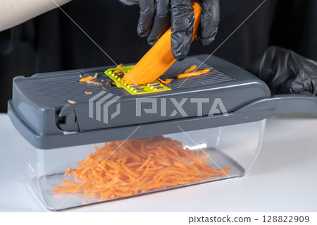 Person's gloved hand expertly grates fresh orange carrot on a modern kitchen slicer, showcasing efficient meal preparation and healthy cooking 128822909