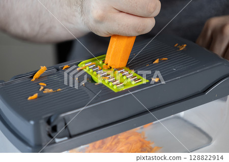 A person's hand skillfully shreds a vibrant orange carrot using a modern kitchen grater, preparing fresh ingredients for healthy meal preparation and home cooking 128822914