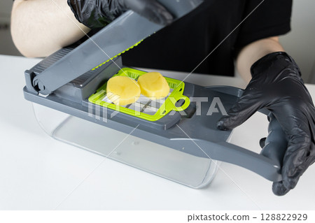 Person in black gloves expertly operates a multi-purpose gray and green vegetable chopper, quickly preparing fresh raw potatoes, symbolizing modern convenient meal preparation and home cooking Person in black gloves expertly operates a multi-purpose gray and green vegetable chopper, quickly preparing fresh raw potatoes, symbolizing modern convenient meal preparation and home cooking 128822929