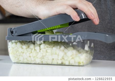 Hand using a modern vegetable chopper to quickly dice white onions, filling the clear container for efficient and convenient home kitchen meal preparation 128822935
