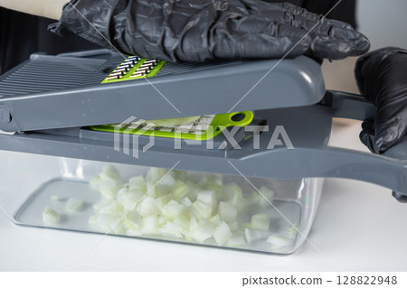 Hands in black gloves operate a modern grey-green vegetable chopper, dicing fresh white onions, illustrating efficient kitchen food preparation 128822948