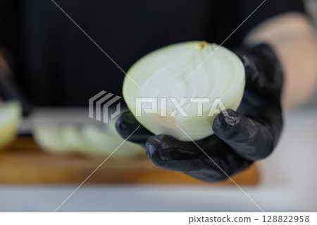 Professional hand in black glove holds freshly cut half white onion, revealing its crisp layers, ideal for cooking, healthy culinary dishes, and kitchen hygiene 128822958