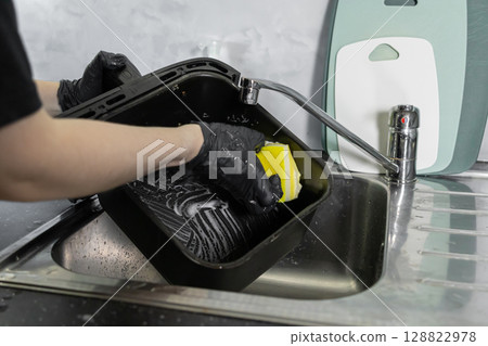 A person wearing protective black gloves diligently cleans an air fryer basket in a kitchen sink, promoting home hygiene and modern appliance care A person wearing protective black gloves diligently cleans an air fryer basket in a kitchen sink, promoting home hygiene and modern appliance care 128822978