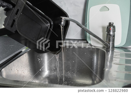 Person wearing black glove pouring dirty water from air fryer basket into a kitchen sink, illustrating effective appliance cleaning 128822980